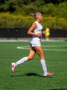 Young female soccer player running on a field, wearing a white jersey and pink cleats, focused on her game.