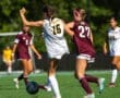 Two female soccer players compete for the ball on a field, one in white and the other in maroon uniforms.