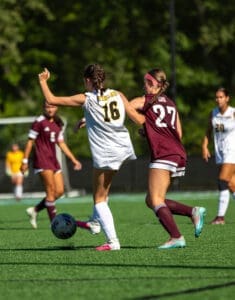 Two female soccer players compete for the ball on a field, one in white and the other in maroon uniforms.