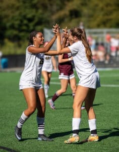 Two female soccer players celebrate with high-fives on the field, showcasing teamwork and sportsmanship on a sunny day.