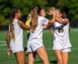 Three female soccer players celebrate a goal with high-fives on the field, wearing matching white uniforms.