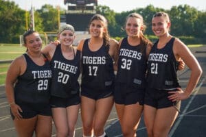 Five female athletes in black uniforms at a sports field, smiling and posing together, showcasing team spirit.