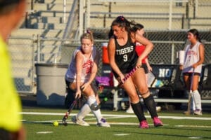 Two young women compete in a field hockey match, showcasing skill and teamwork on the turf.