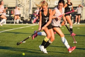 Two girls actively compete in field hockey, dribbling a ball on a sunny day while wearing team jerseys and focusing intently.