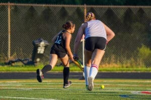 Two young athletes compete in field hockey on a grass field, actively pursuing the ball under the sun.