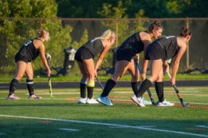 Four field hockey players in black uniforms prepare for practice on a sunny day, focusing on their techniques on the turf.