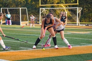 Two field hockey players compete for the ball on a turf field, with the goalie and teammates in the background.