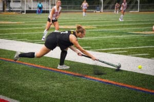 Field hockey player lunges to strike a ball during a competitive match on a synthetic turf field. Team in background.