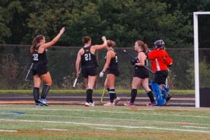 Field hockey players celebrating together on the field, wearing black uniforms, with a goalie in orange gear nearby.