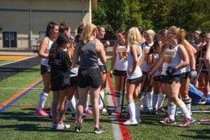 Field hockey team huddles together, strategizing before a game on a sunny day. Team spirit and determination are evident.