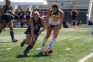 Two female field hockey players compete for the ball on a sunny day, showcasing athleticism on the turf field.