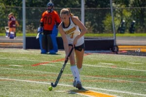 Field hockey player in a Tigers uniform dribbling a ball on a sunny day, with goalkeeper in the background.