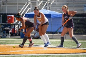 Three field hockey players compete on the turf, focused on the ball, with spectators and equipment in the background.