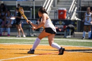 Field hockey player prepares to strike the ball on a bright orange turf with teammates in the background.