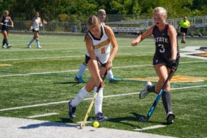 Two high school girls compete in field hockey on a sunny day, focusing on the ball on a synthetic turf field.