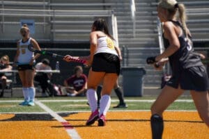 Field hockey players in action during a match on a vibrant orange turf field. Focus on athlete with pink stick.