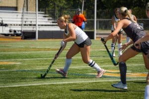 Field hockey player dribbling a ball on the turf during a sunny practice session with teammates in the background.