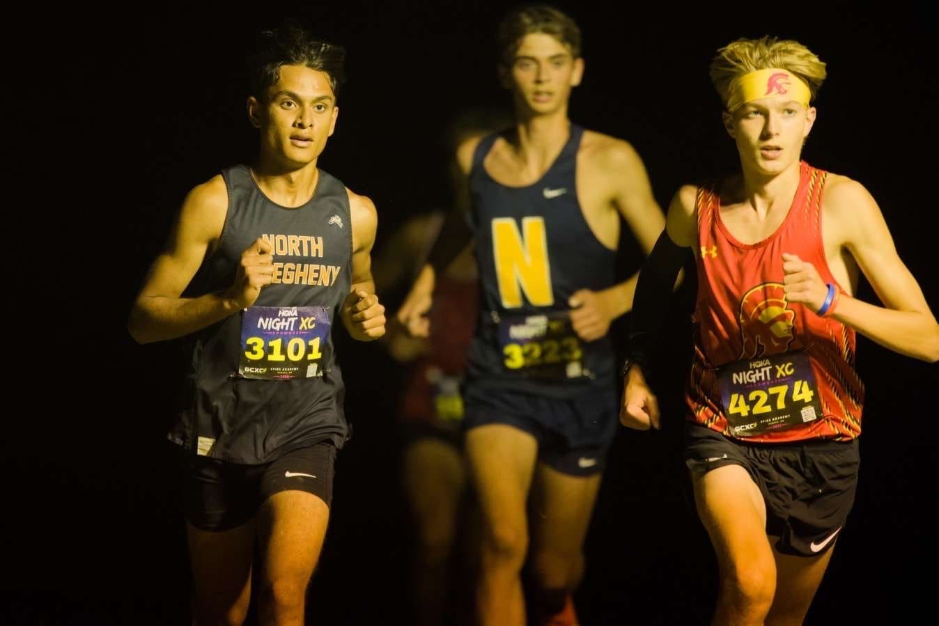 Three young male athletes race in a night cross country event, wearing numbered jerseys and focused expressions.