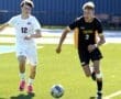 Two high school soccer players compete for the ball on a sunny, green field during an intense match.