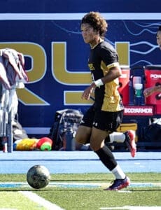 Player in a black and gold soccer uniform dribbling the ball during a game, focused on the field ahead.