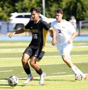 High school soccer players in action, one in a black jersey dribbling the ball, another close behind in white.
