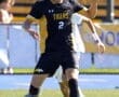 High school soccer player in black jersey dribbles a ball on the field during a game against an opponent.