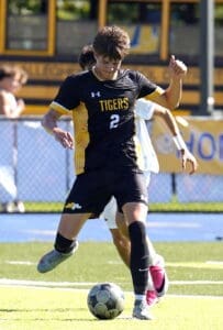 High school soccer player in black jersey dribbles a ball on the field during a game against an opponent.
