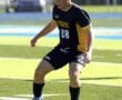 Young male soccer player in black uniform dribbling a ball on a sunny field during a game, focused on the action.