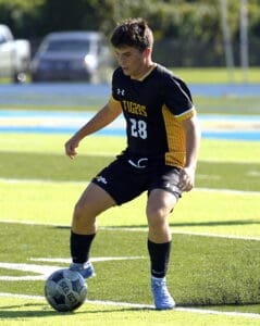 Young male soccer player in black uniform dribbling a ball on a sunny field during a game, focused on the action.