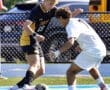 Two soccer players in a competitive match, one in black and yellow, the other in white, attempting to gain control of the ball.