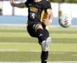 Soccer player in black uniform kicking the ball on a grassy field during an outdoor game, showcasing athleticism.