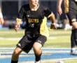 High school soccer player in a black and gold uniform dribbles the ball on a field during an intense match.