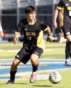High school soccer player in a black and gold uniform dribbles the ball on a field during an intense match.