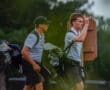 Two young golfers walking on a course, one gesturing with his hand, both carrying golf bags.