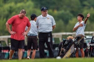 Four individuals at a golf course including a coach and players, discussing strategy near golf bags and equipment.