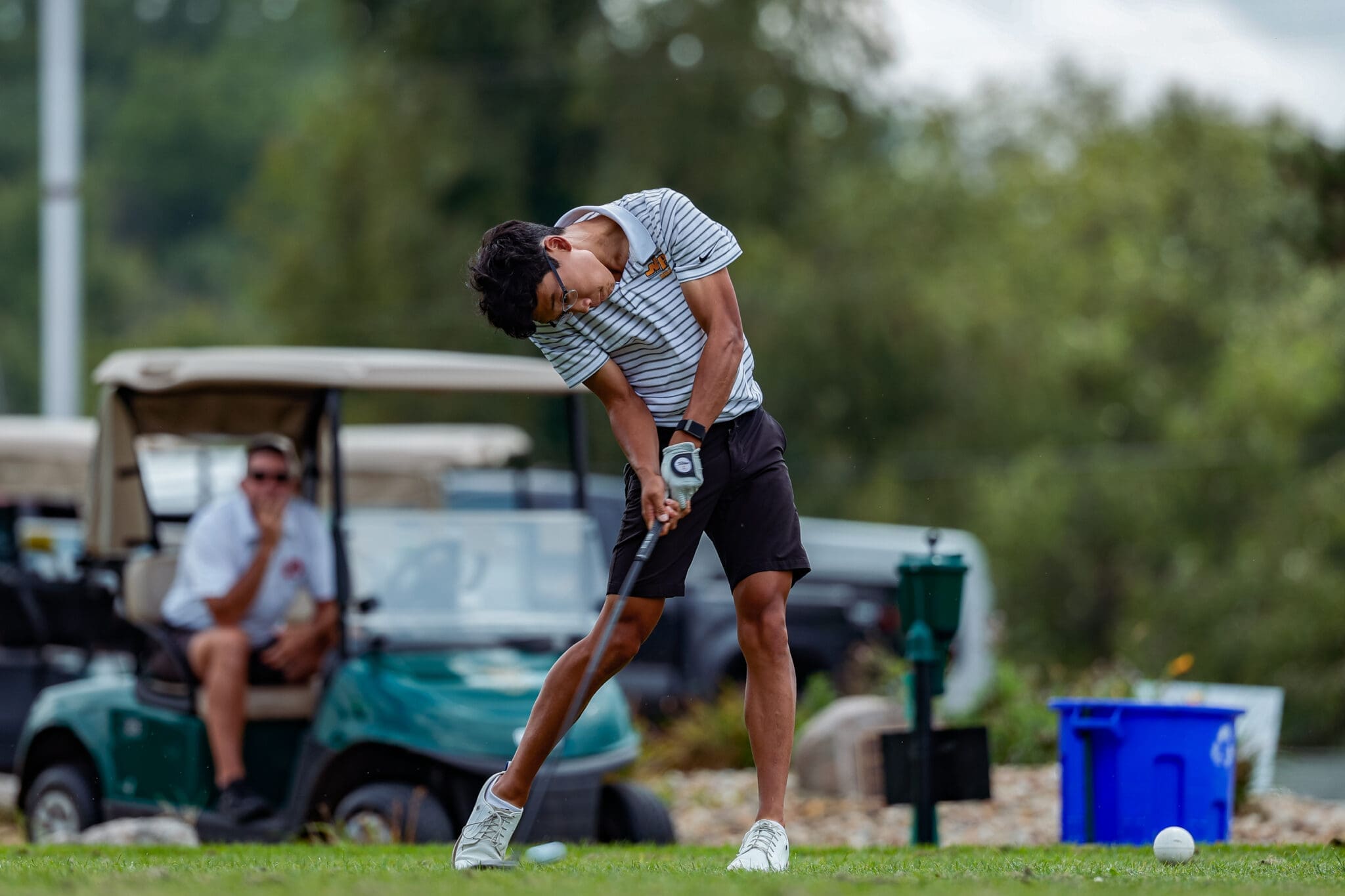 A young golfer swings his club on the course, with a golf cart and fellow player in the background.