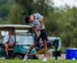 A young golfer swings a club on the course, with a golf cart and a spectator in the background.