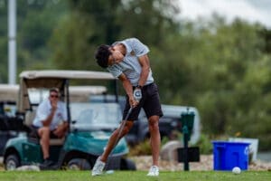 A young golfer swings a club on the course, with a golf cart and a spectator in the background.
