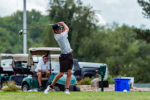 Young golfer preparing to hit a drive, with golf carts in the background on a lush green course.