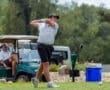 Young golfer swings a driver on the course, with golf carts and a spectator in the background.