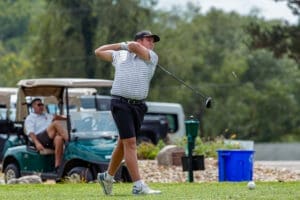 Young golfer swings a driver on the course, with golf carts and a spectator in the background.