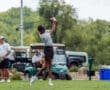 A young golfer swings a driver on the course, with spectators and golf carts in the background.