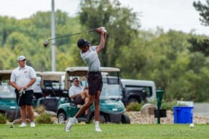 A young golfer swings a driver on the course, with spectators and golf carts in the background.