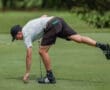 A golfer in a striped shirt leans down to retrieve his ball from the green on a sunny day.