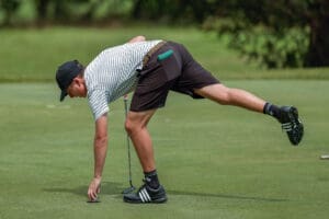 A golfer in a striped shirt leans down to retrieve his ball from the green on a sunny day.