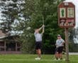 A young golfer swings at a tee while two friends watch, with a course sign displaying hole details in the background.