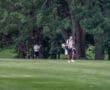 A golfer carrying a bag walks on a lush green course, with another player checking their scorecard in the background.