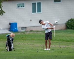 Young golfer swinging a club on a lush green course, with golf clubs and a plush toy in the background.