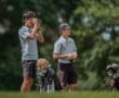 Two young golfers on a course; one aims a rangefinder while the other holds a scorecard. Golf equipment is nearby.