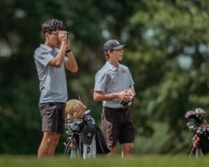 Two young golfers on a course; one aims a rangefinder while the other holds a scorecard. Golf equipment is nearby.